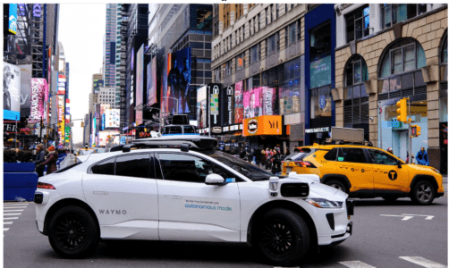 A self-driving car navigates a busy street filled with other vehicles and pedestrians.