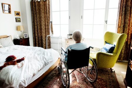 An elderly woman in a wheelchair faces a bright window in a cozy bedroom with a bed, nightstand, green armchair, and patterned rug illuminated by natural light.