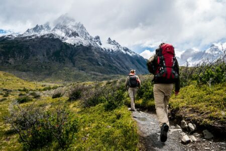 Two hikers with backpacks walking along a mountain trail surrounded by lush greenery and rocky terrain.