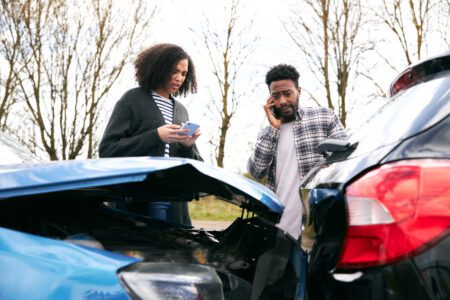 A man and woman converse on the phone beside a damaged car, appearing concerned about the situation.