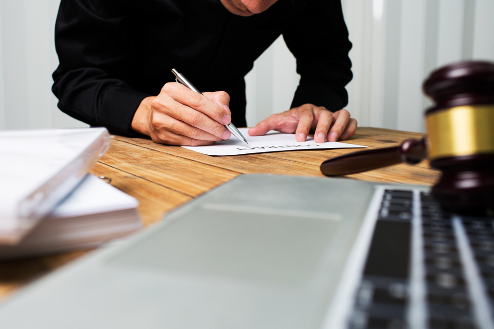 A man in a black shirt writes on paper at a wooden desk, with a laptop and a gavel nearby.
