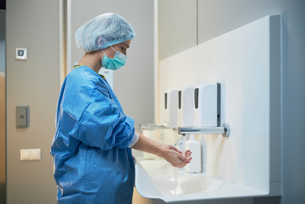 Healthcare worker in scrubs, surgical cap, and mask washing hands at a sink