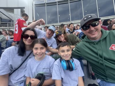 Woman, girl, boy, and man taking a selfie in the stands at a sporting event, with 2 other people cheering behind them and text on the shirts: West Point