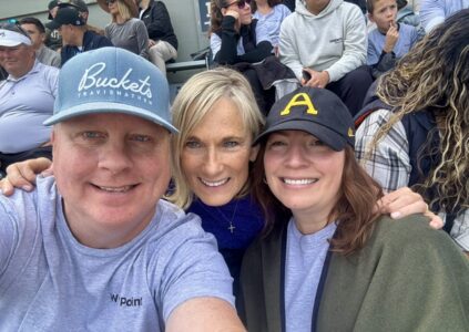 Man in a hat with text: Buckets TravisMathew, and 2 women taking a selfie in the stands at a sporting event