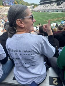 Woman giving a thumbs-up in the stands of a football stadium, wearing a shirt with text: Finkelstein & Partners The Injury Attorneys, Fine, Olin & Anderman Attorneys for Workers, Diller Law, Legal Ally