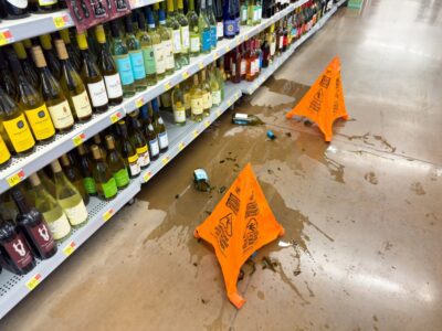 Spilled wine and broken bottles on a store floor next to wine shelves, marked by cones with text: Caution Wet Floor