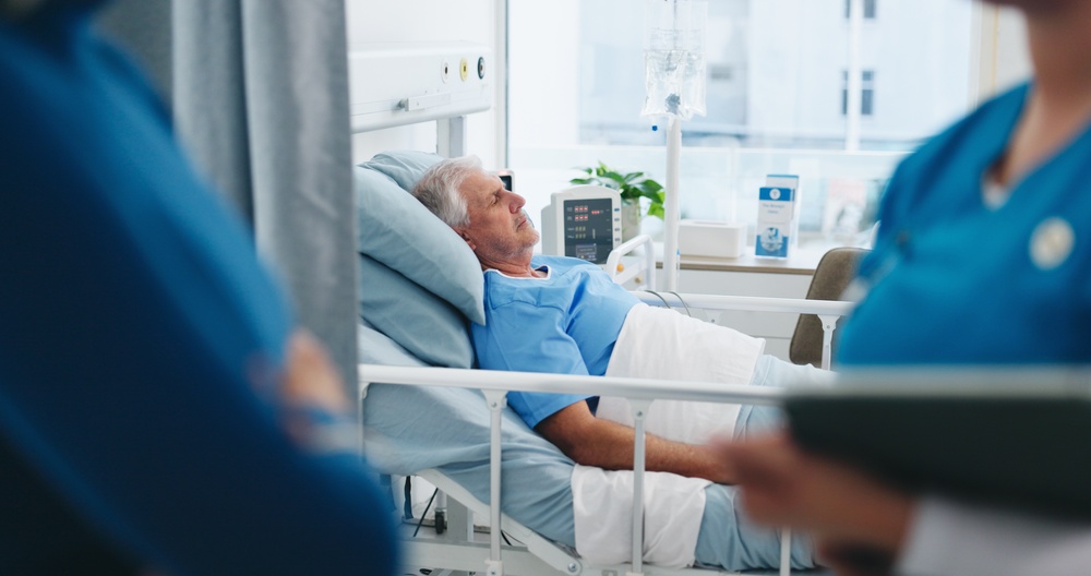Elderly man asleep in a hospital bed as medical staff stand nearby